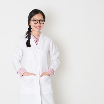 Portrait of young Asian female scientist with lab coat standing on plain background.