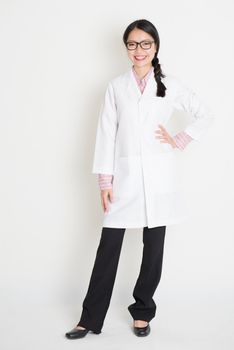 Full length portrait of young Asian female scientist with lab coat standing on plain background.