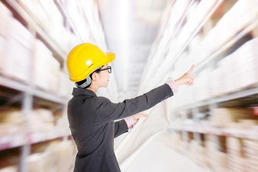 Young Asian female store manager with hard hat counting stock at warehouse. Shelves with goods at background.