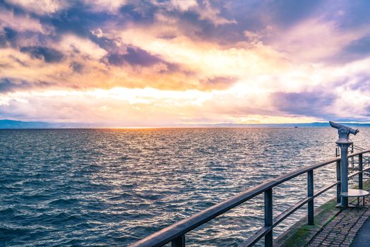 Landscape with a colorful sunset over the Bodensee lake and its shore, equipped with tourists binoculars and metal railing for safety.
