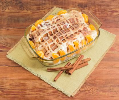 Sweet pancakes filled with cottage cheese with sour cream and sprinkled with cinnamon powder and mandarin orange segments in the rectangular glass roasting pan and cinnamon sticks on cloth napkin on old wooden surface
