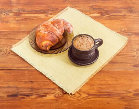 Freshly brewed coffee latte in a black ceramic cup and croissant on a glass saucer on a placemat on an old wooden surface
