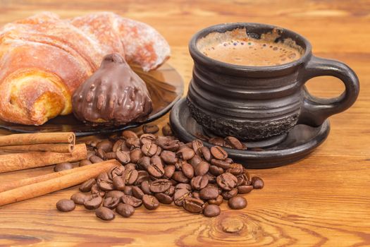 Freshly brewed coffee latte in a black ceramic cup closeup on the background of roasted coffee beans, cinnamon sticks, chocolate truffle and croissant on an old wooden surface
