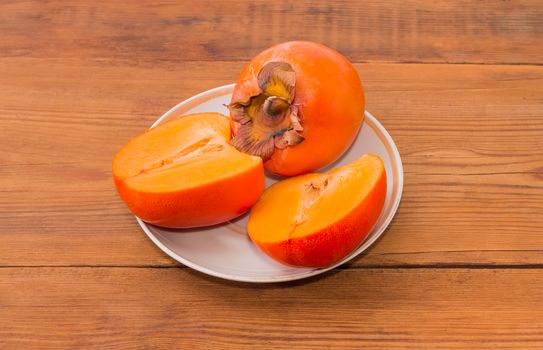 One whole ripe fresh persimmon and one cut in half of a persimmon on saucer on a wooden surface
