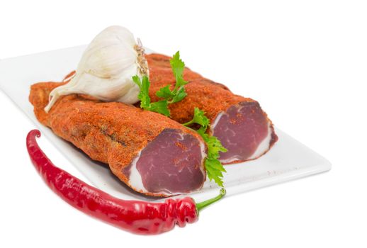 Two pieces of dried pork tenderloin on a white dish, garlic, one fresh chili and twig of parsley closeup on a light background 
