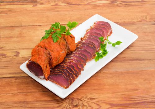 One piece and sliced dried pork tenderloin with twigs of parsley on a white rectangular dish on a wooden surface
