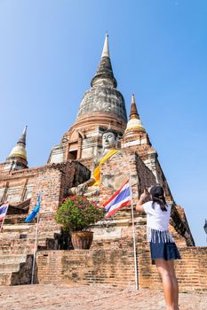 Tourist teenage girl standing take a photo of buddha statue and ancient pagoda on blue sky background at Wat Yai Chaimongkol temple in Phra Nakhon Si Ayutthaya Historical Park, Thailand