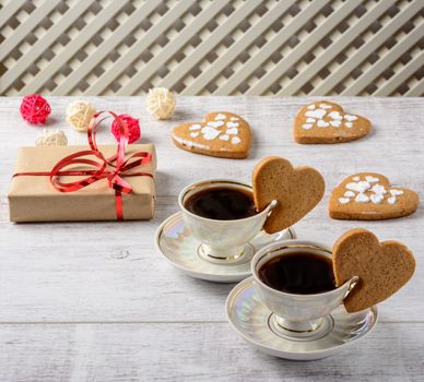 Box with a present to the Valentine's Day cookies in the shape of a heart and cups of black coffee on the white table