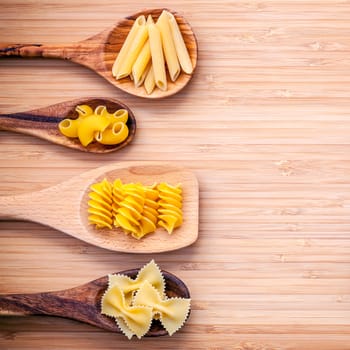 Italian foods concept and menu design. Various kind of Pasta Elbow Macaroni ,Farfalle ,Fusilli  and Penne in wooden spoons setup on bamboo cutting board flat lay and copy space.