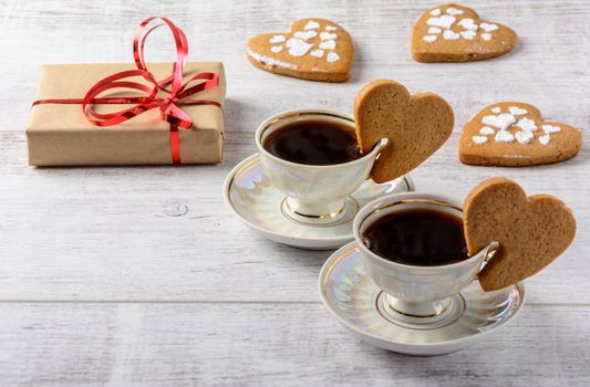 Box with a present to the Valentine's Day cookies in the shape of a heart and cups of black coffee on the white table