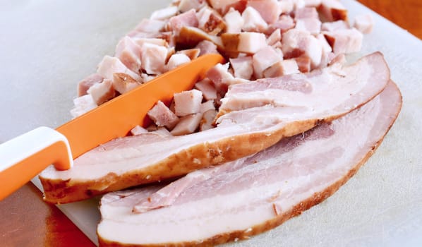 Closeup of bacon slices and cubes and orange knife on cutting board.