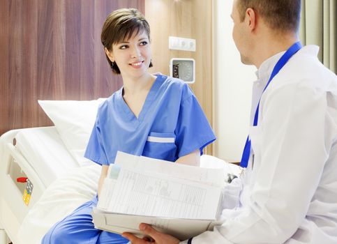 A happy young female patient sitting on a modern electrical hospital bed, smiling at a doctor with files in hands.