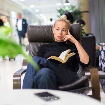 Thougtful woman sitting by wooden table and reading book in public library.