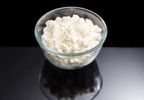 Transparent glass bowl with fresh cottage cheese on a dark background
