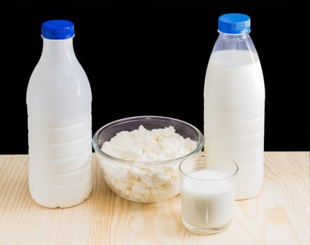 One plastic bottle with milk, one plastic bottle with fermented milk drink, glass bowl with cottage cheese, glass with milk on a wooden surface on a dark background
