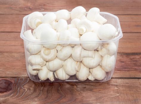 Fresh uncooked button mushrooms in a transparent plastic tray on a dark wooden surface
