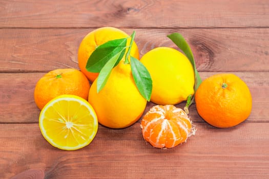 Several fresh whole mandarins, oranges and lemons with twigs and leaves, one lemon cut in half, one peeled mandarin on a dark wooden surface
