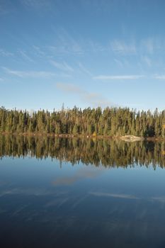 Dead calm lake in early morning with reflections of conifers and wispy cirrus clouds