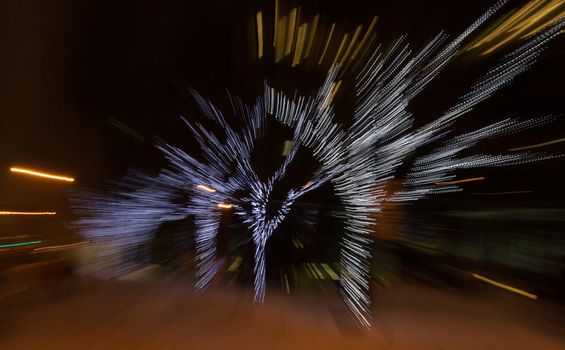 Four Christmas holiday light covered trees are blurred into a dynamic, bright night time image.  Office building lights and streetlights are also intentionally blurred in this slightly distubing, fast-moving image.