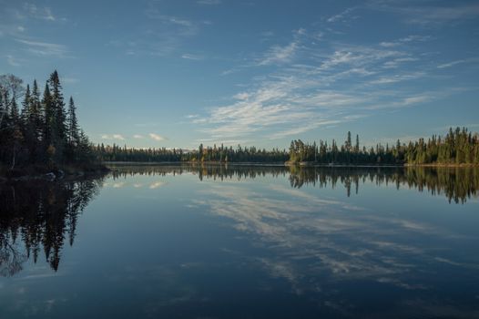 Dead calm lake in early morning with reflections of conifers and wispy cirrus clouds