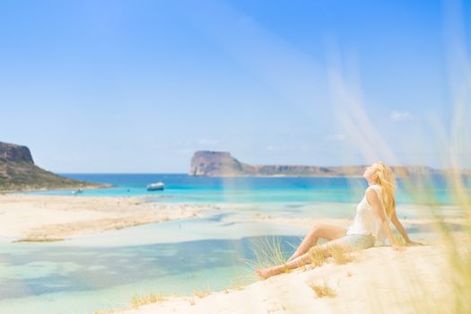 Relaxed woman enjoying sun, freedom and life an a beautiful sandy beach of Balos in Greece. Young lady feeling free, relaxed and happy. Vacations, freedom, happiness, enjoyment and well being.
