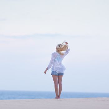 Meditative, sensual blonde woman wearing white loose casual shirt on vacations, looking at horizon, enjoying peaceful early morning.