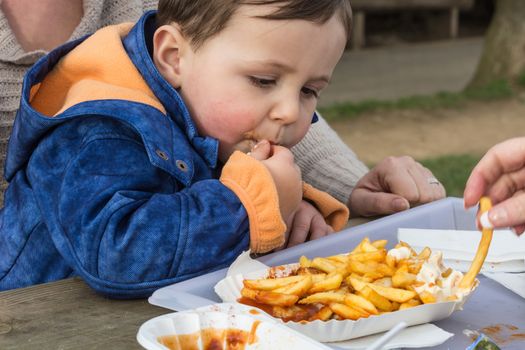 Child eats sausage with french fries from a paper plate.