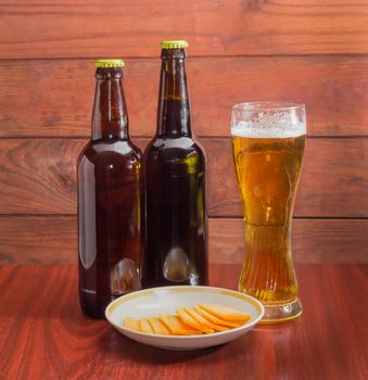 Glass of lager beer, two bottles of beer and several slices of cheese on saucer on wooden table against the backdrop of dark wooden planks
