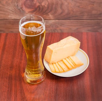 Beer glass with lager beer, one piece and several thin slices of hard cheese on saucer on wooden table against the backdrop of dark wooden planks
