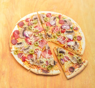 Sliced cooked round pizza with various sausages, button mushrooms and olives on wooden cutting board on a light background
