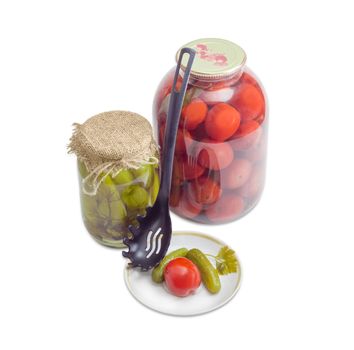 Two pickled cucumbers and one pickled tomato on a saucer, two various glass jars with pickled cucumbers and tomatoes and black plastic slotted spoon on a light background

