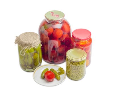 Two pickled cucumbers and one pickled tomato on a saucer and pickled cucumbers, tomatoes, bell pepper and green peas in glass jars on a light background
