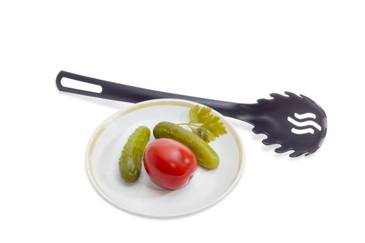 Two pickled cucumbers and one pickled tomato on a saucer and black plastic slotted spoon on a light background

