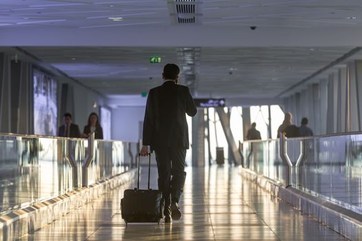 Businessman dragging a small carry on luggage suitcase at airport corridor walking to departure gates.