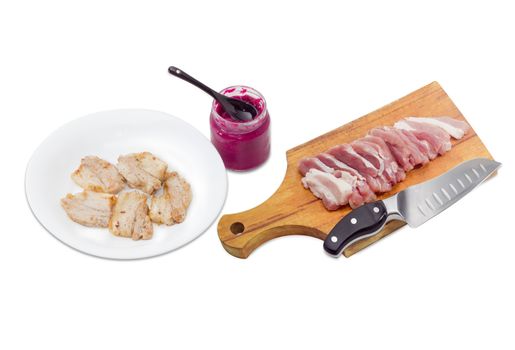 Several slices of fried pork on white dish,  small glass jar of horseradish sauce,  sliced uncooked pork belly and kitchen knife on a wooden cutting board on a light background
