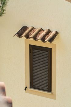 Window with brown closed shutters. Rain protection from roof tiles in a yellow painted house wall.