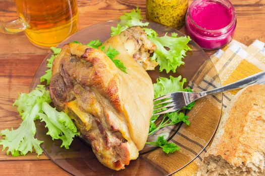 Baked ham hock with lettuce and parsley and fork on a glass dish on the background of a beer and condiments closeup
