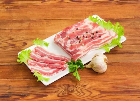 Uncooked partly sliced streaky pork belly bacon with spice, garlic and greens on a rectangular dish on a old wooden surface
