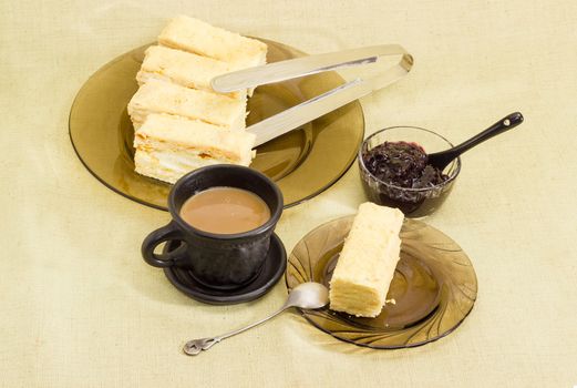 Piece of layered sponge cake on a glass saucer with spoon, several pieces of the rest of the cake on glass dish, coffee with milk in black cup and small glass bowl with jam on a cloth surface
