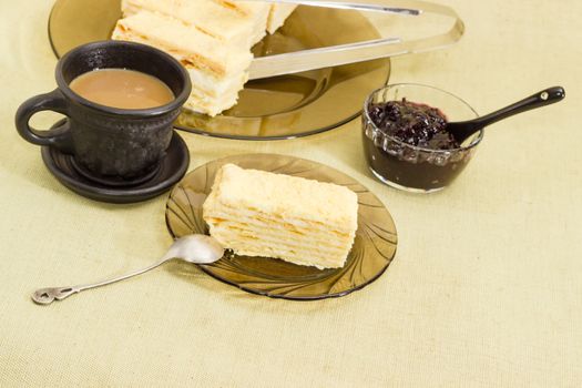 Piece of layered sponge cake on a glass saucer with spoon on the background of the rest of the cake, coffee with milk in a black cup and jam on a cloth surface
