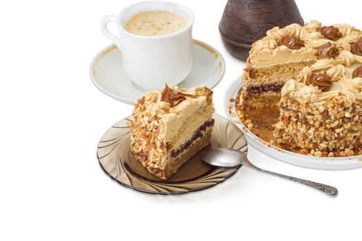 Partly sliced layered sponge cake, decorated with butter cream, caramelized condensed milk and nuts on background of coffee cup and coffee pot on a light background

