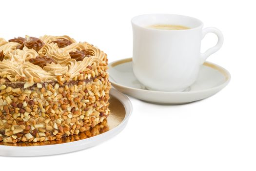 Fragment of a round sponge cake, decorated with butter cream and caramelized condensed milk, sprinkled with grated nuts on a background of white cup with coffee with cream on a light background
