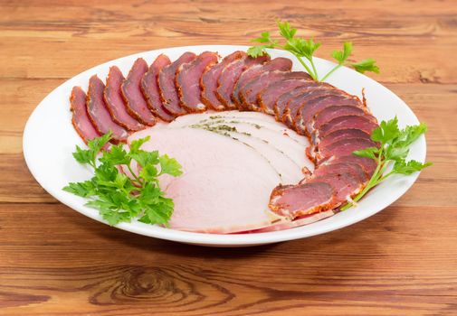 Sliced dried pork tenderloin and ham with twigs of parsley on a white dish on a wooden surface
