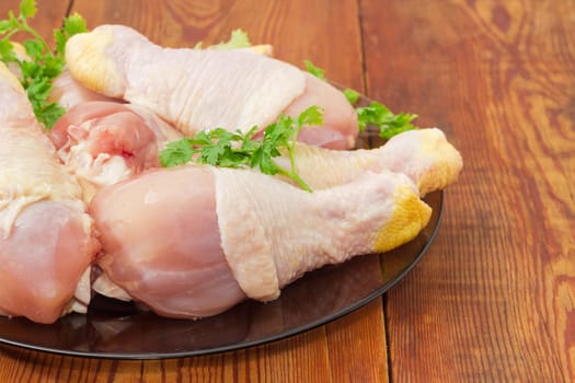 Fragment of a glass dish with uncooked chicken legs and twigs of parsley closeup on an old wooden surface
