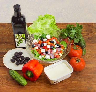 Greek salad in a glass salad bowl among ingredients for its cooking on an old wooden surface
