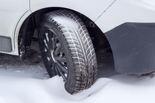 Front wheel of the white car with a snow tire, covered partly mud and partly snow closeup during snowfall
