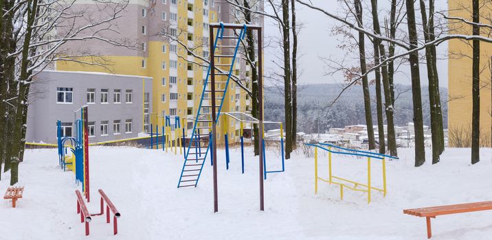 Panorama of outdoor gym in winter public park coverd snow on the background of modern multistory apartment complex
