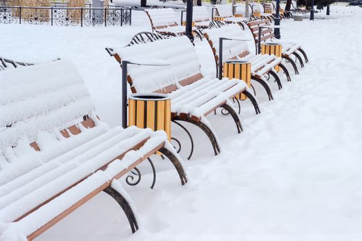 Rows of park benches, made of forged steel frame and wooden planks and covered with snow in a public garden on a cloudy day
