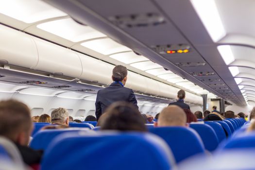 Interior of airplane with passengers on seats and stewardess walking the aisle.