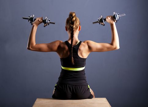 Fit young woman doing shoulder raises with dumbbells, on the wood box